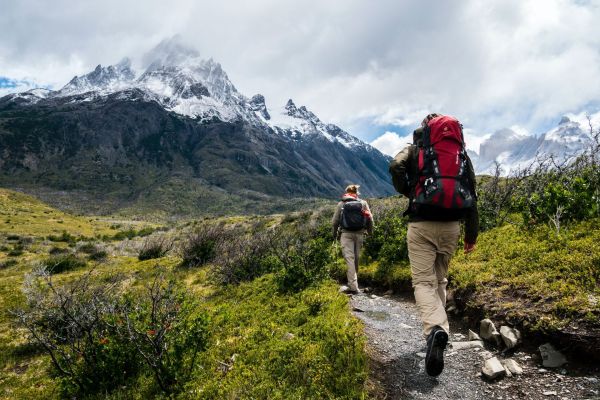 2 Personen mit Rucksäcken Wandern auf einem Weg. Im Hintergrund sind Schneebedeckte Berge zu sehen.